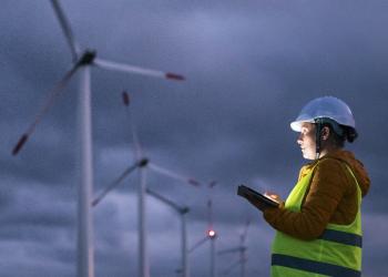 Construction worker types on a tablet while standing near six wind turbines